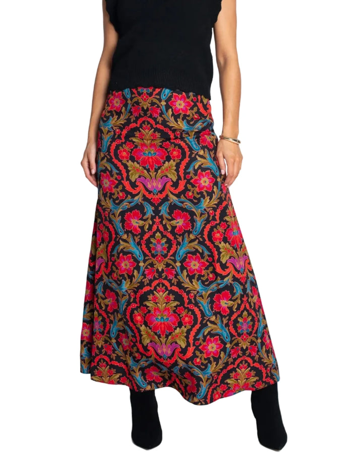 A model wearing a sleeveless black turtleneck top and a long, full A-line skirt with a vibrant red, blue, and gold damask pattern, standing against a plain white studio background.