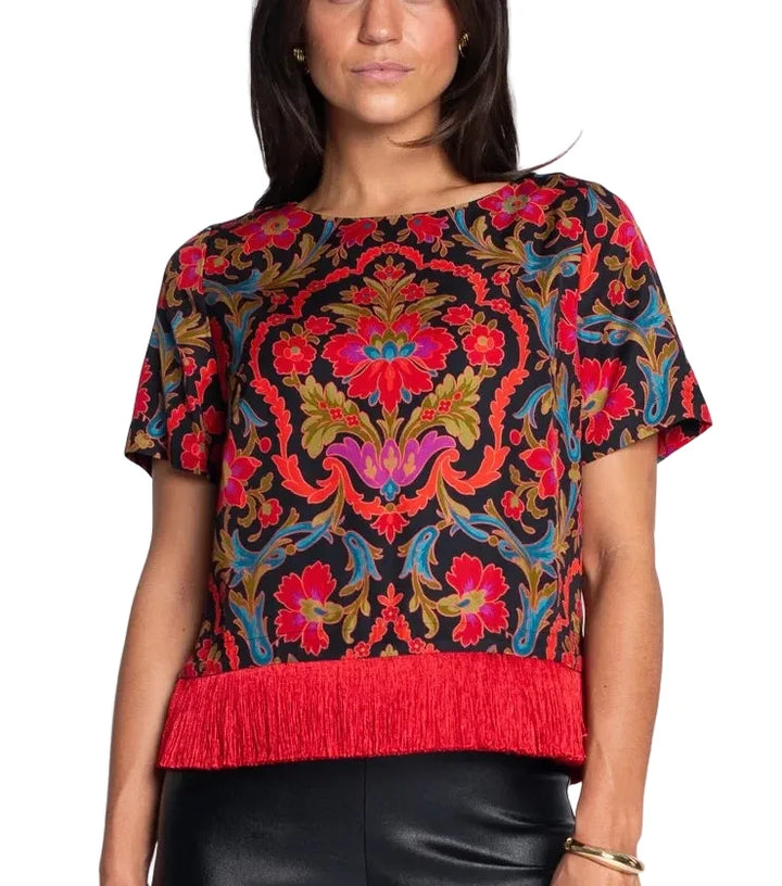 A woman wearing a short-sleeve bohemian-style top with a vibrant red fringe hem, featuring a multicolor floral and paisley pattern on a black background.