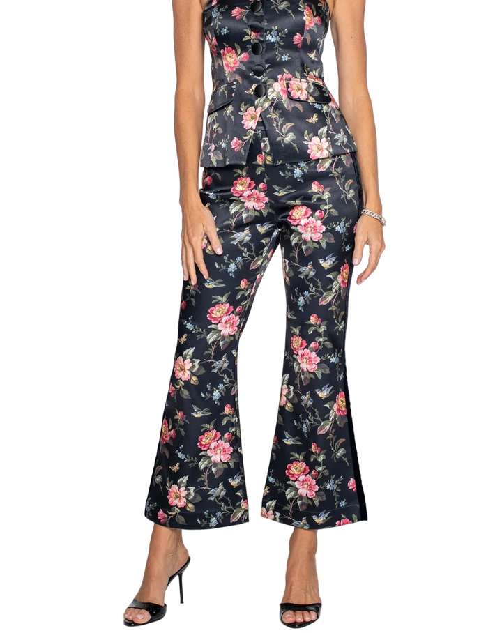 A woman modeling a strapless black satin jumpsuit with a pink and green floral print, featuring a peplum waist and wide-leg cropped pants, set against a plain light gray background.