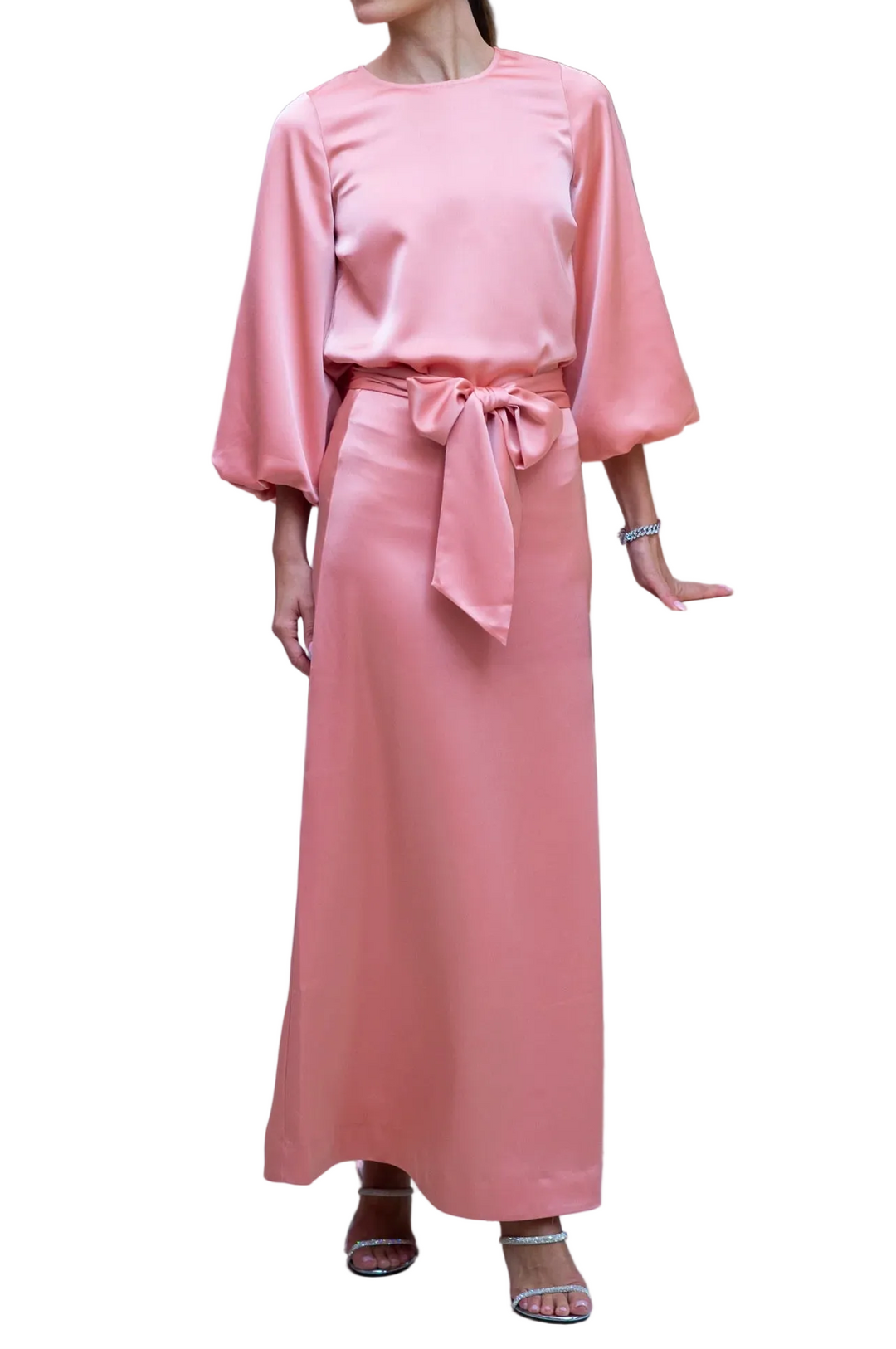 A woman models a dusty pink two-piece set consisting of a long-sleeve satin top and a matching maxi skirt with a tie waist, set in an elegant indoor environment.