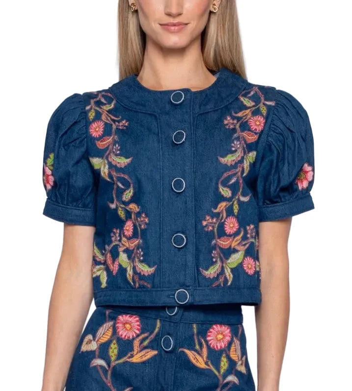 A woman modeling a dark blue denim-like cropped top with puff sleeves and colorful floral embroidery, paired with matching bottoms against a plain light gray studio background.