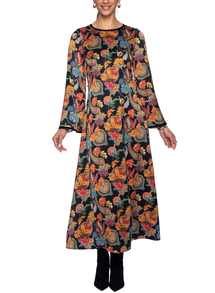 A woman modeling a long-sleeved midi dress with a vibrant multicolor paisley and floral print on a black background, paired with black ankle boots, against a light gray studio backdrop.