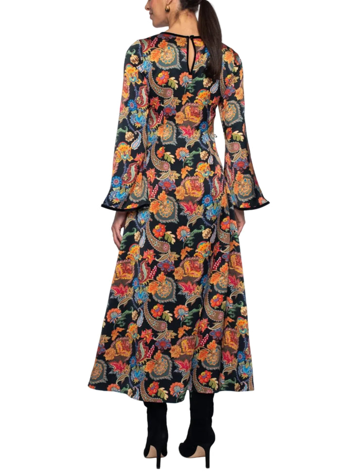 A woman viewed from the back wearing a long-sleeved maxi dress with a vibrant multicolor paisley and floral print on a black base, paired with black boots against a white studio background.