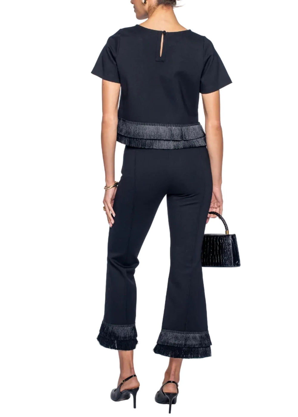 A woman shown from the back wearing a black short-sleeve top and matching black flared pants with textured fringe trim at the waist and cuffs, paired with black heels and holding a small black handbag against a light gray studio background.