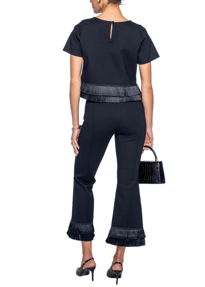 A woman shown from the back wearing a black short-sleeve top and matching black flared pants with textured fringe trim at the waist and cuffs, paired with black heels and holding a small black handbag against a light gray studio background.