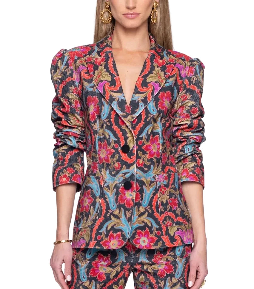 A woman modeling a matching two-piece suit with a vibrant, ornate baroque-style floral and scroll pattern in red, pink, blue, and gold on a dark background, set against a plain white studio backdrop.