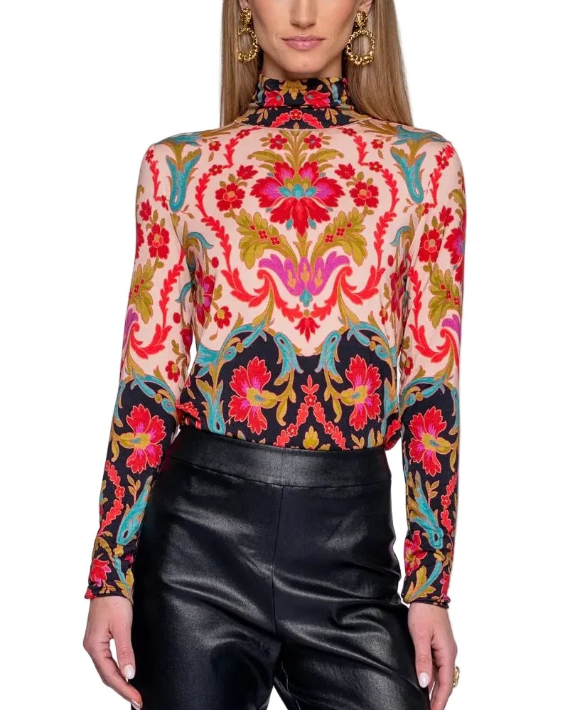 A woman modeling a long-sleeve mock neck top with a vibrant, multicolor baroque-style floral and ornamental pattern on a cream background, paired with black leather pants against a light gray studio background.