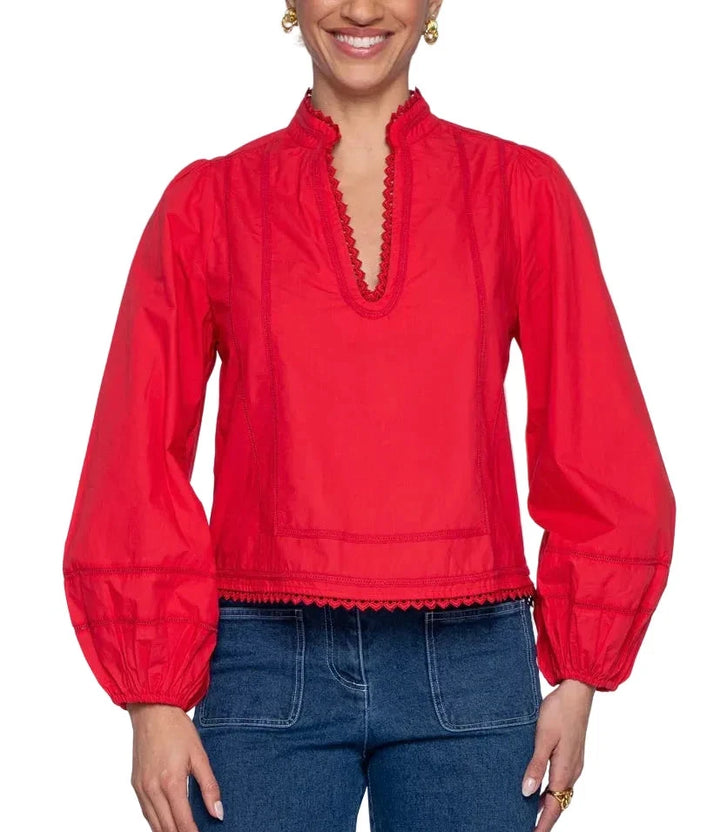 A woman wearing a bright red long-sleeve blouse with a high gathered collar, scalloped trim, and voluminous sleeves, paired with blue jeans against a light gray studio background.