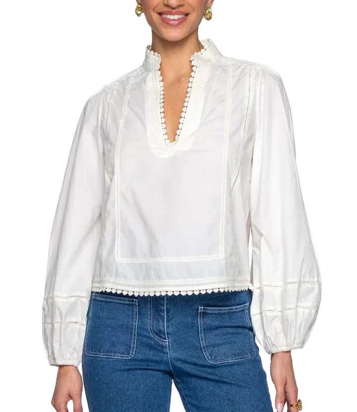 A woman wearing a white long-sleeve blouse with a high ruffled collar, decorative trim, and gathered cuffs, paired with blue jeans, against a plain white studio background.