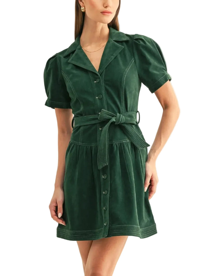 A woman modeling a dark green corduroy mini dress with puff sleeves, a button-up front, and a matching tie belt at the waist, set against a plain light background.
