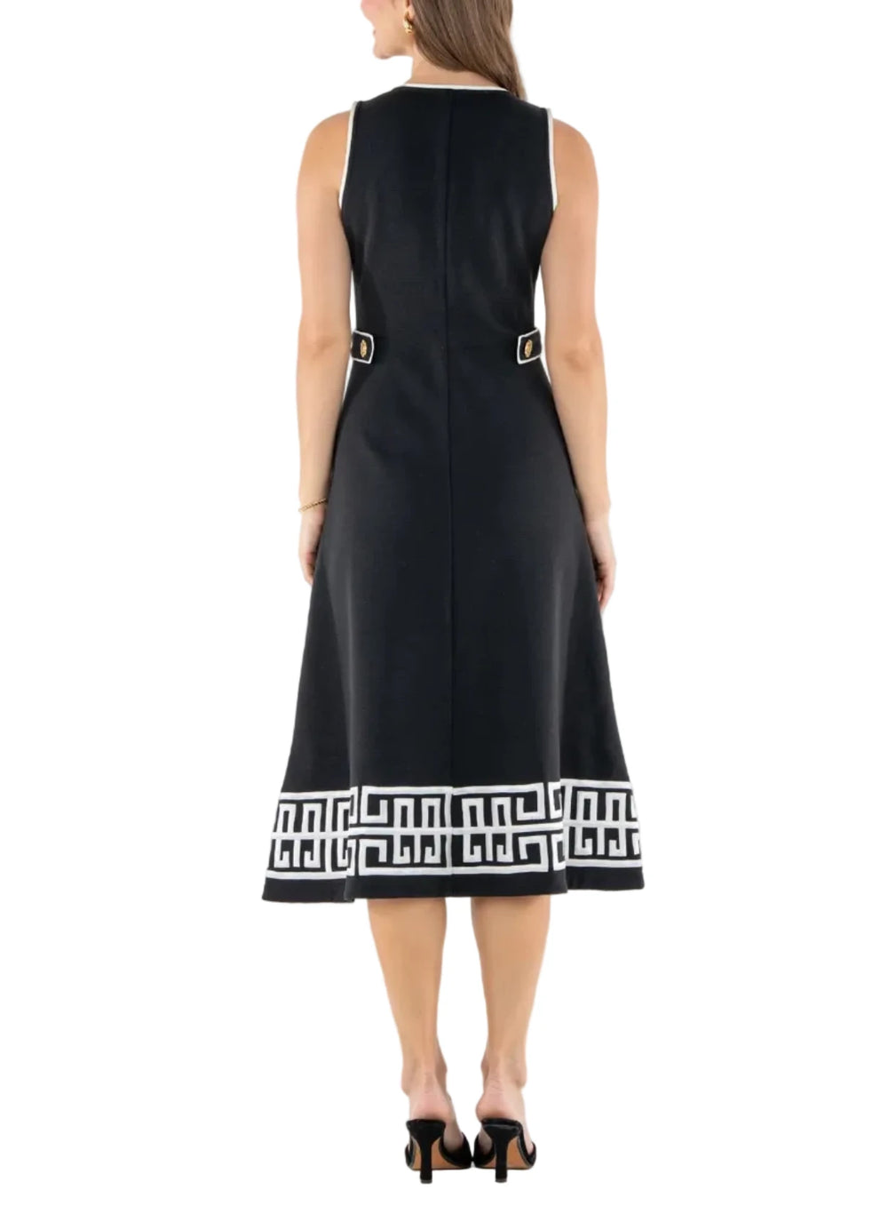 A woman viewed from the back wearing a sleeveless black midi dress with a white geometric border at the hem and small gold-tone buckle details near the waist, photographed against a plain white studio background.