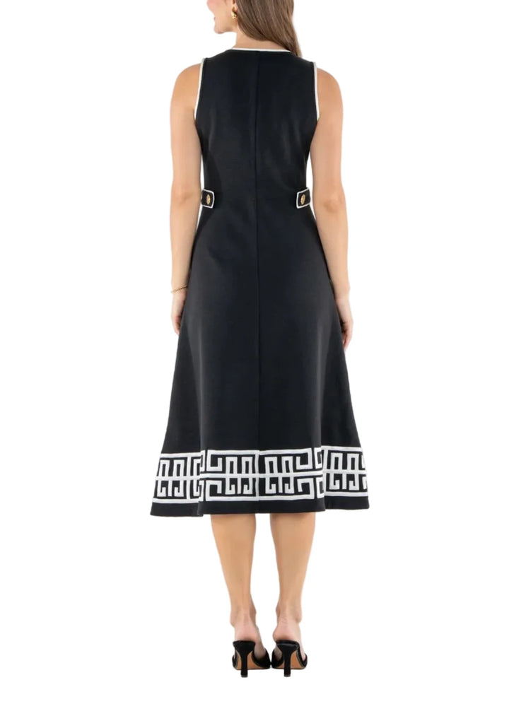 A woman viewed from the back wearing a sleeveless black midi dress with a white geometric border at the hem and small gold-tone buckle details near the waist, photographed against a plain white studio background.