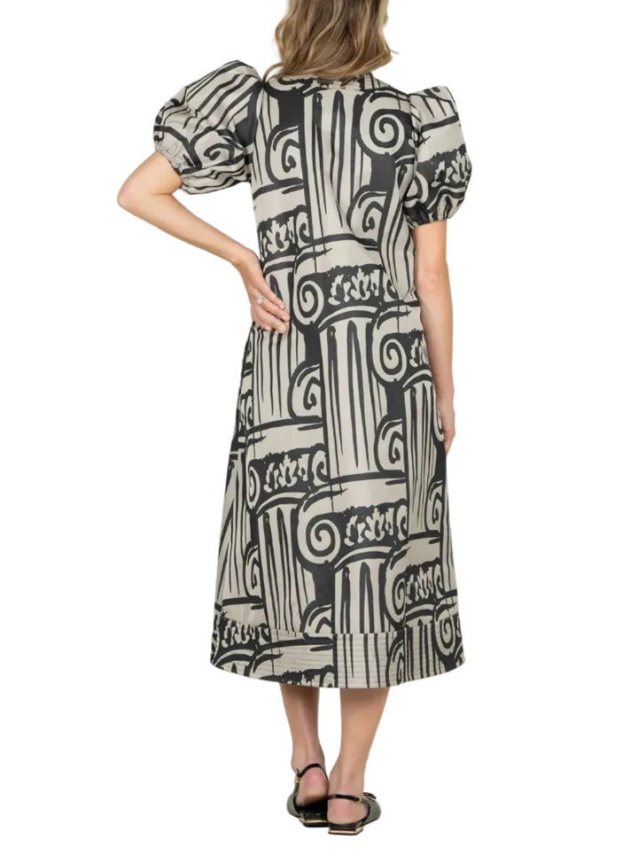 A woman viewed from the back wearing a knee-length dress with puff sleeves and an abstract black, white, and gray column-like print, standing against a plain light gray studio background.
