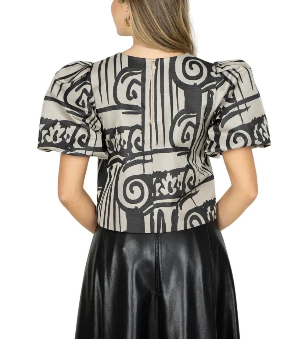 A woman viewed from the back wearing a short-sleeve puff-sleeve top with a black and beige geometric pattern, paired with a black skirt, against a light gray studio background.