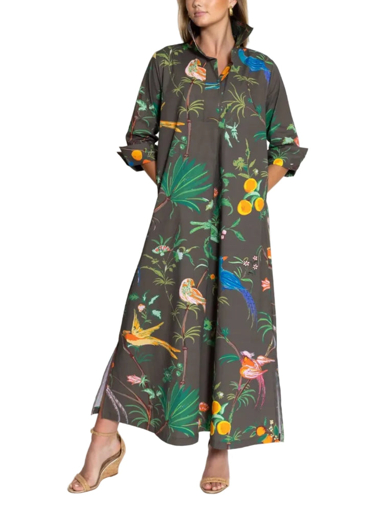 A woman modeling a dark brown maxi dress with a vibrant tropical print featuring green palm leaves, colorful birds, and orange fruits, set against a plain white studio background.