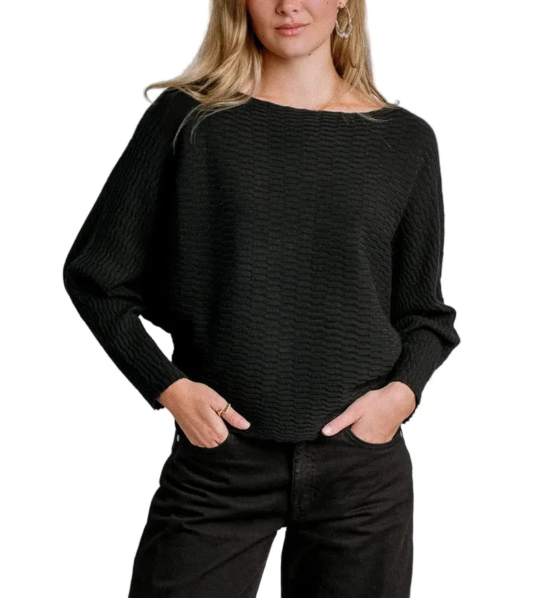 A woman modeling a black textured knit sweater with a wide boat neckline and long sleeves, paired with black pants, against a light gray studio background.