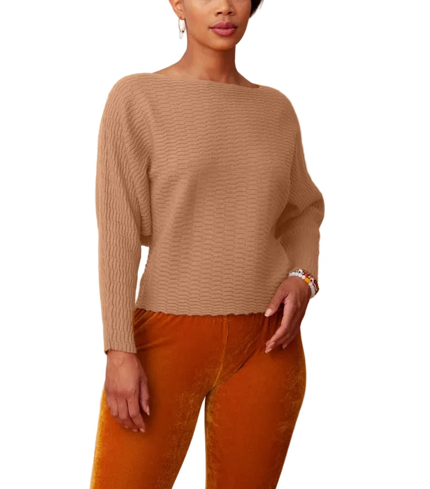 A woman modeling a beige ribbed knit sweater with a boat neckline, paired with orange velvet pants, against a textured beige backdrop.