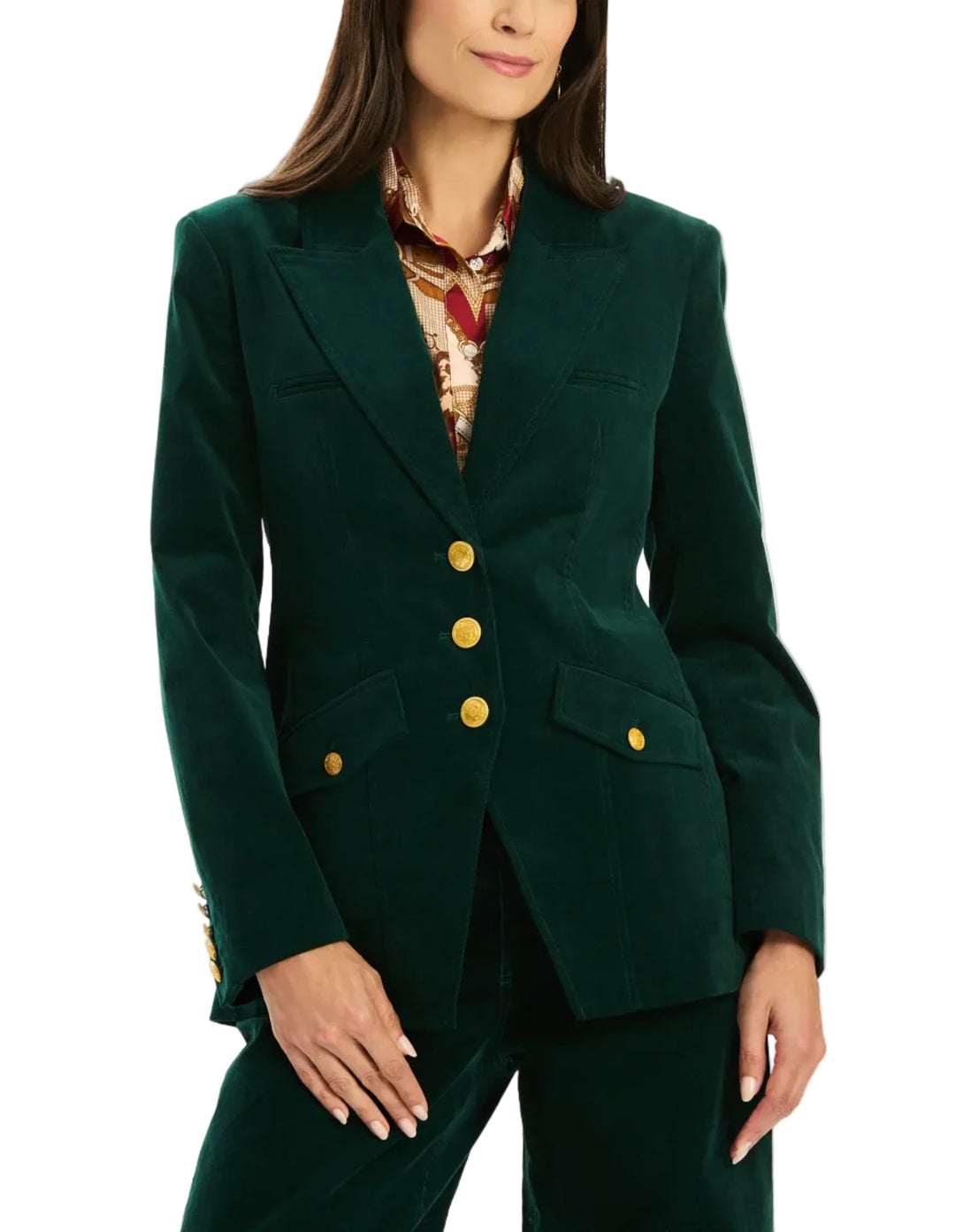 A woman modeling a dark green velvet blazer with gold buttons, paired with matching green trousers and a patterned scarf, against a plain white studio background.