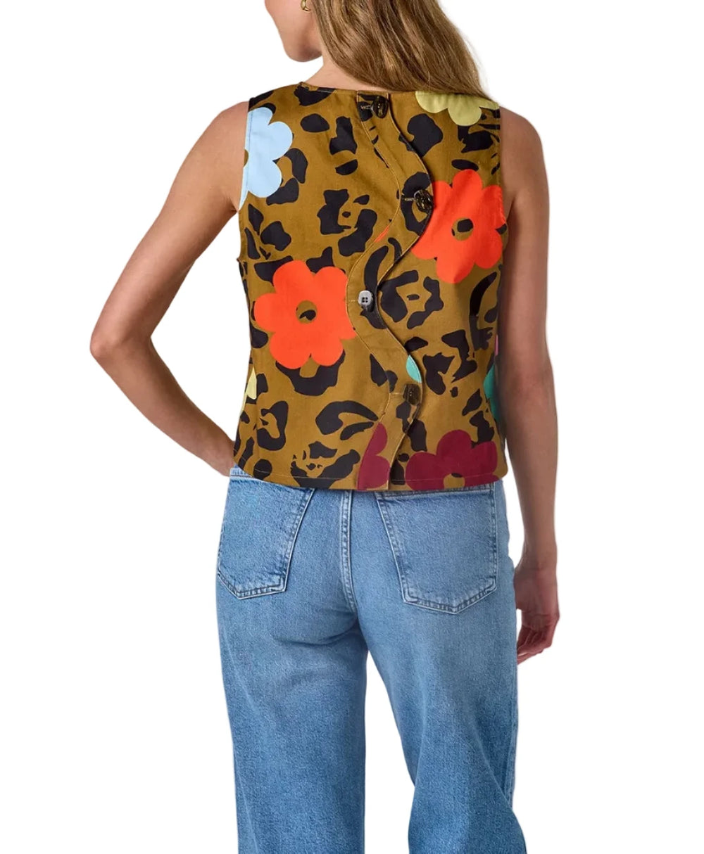 A woman shown from the back wearing a sleeveless cropped top with a mustard yellow leopard print base and large colorful floral motifs, paired with light blue jeans, against a plain light gray studio background.