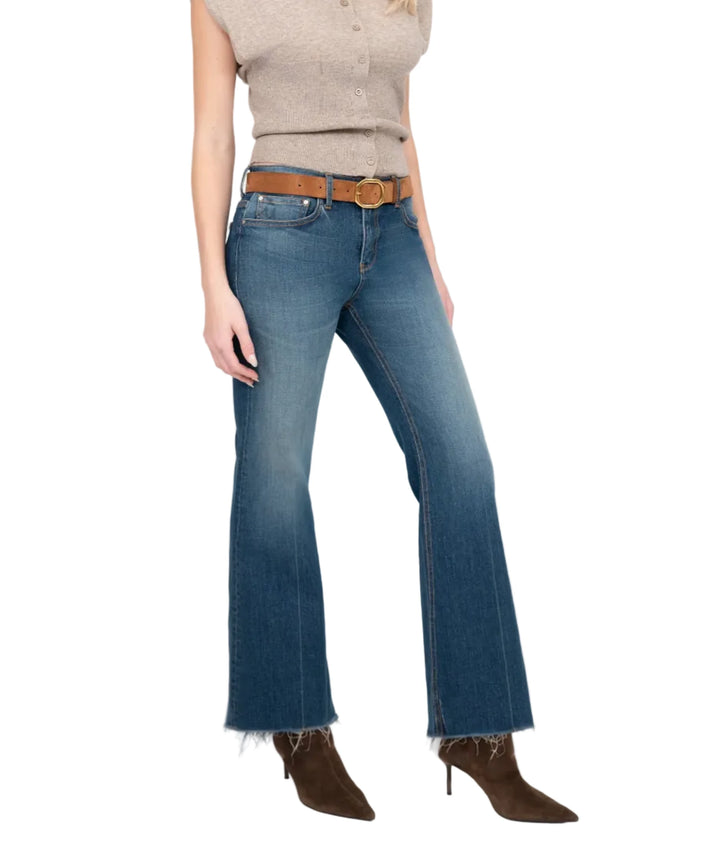 A woman modeling a beige sleeveless knit top and blue flare jeans with a brown belt and brown heeled boots, photographed against a light gray studio background.