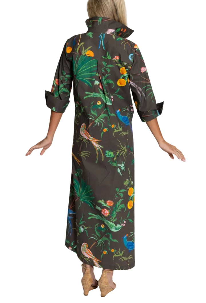 A woman shown from the back wearing a dark brown high-neck maxi shirt dress with a colorful tropical print of birds, foliage, and fruit, paired with tan boots against a light gray studio background.