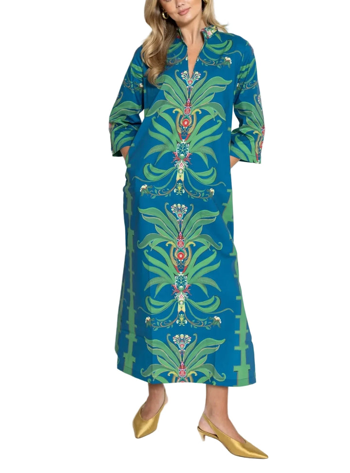 A woman modeling a long-sleeved maxi dress with a vibrant green and teal botanical pattern, paired with gold mule heels, against a plain white studio background.