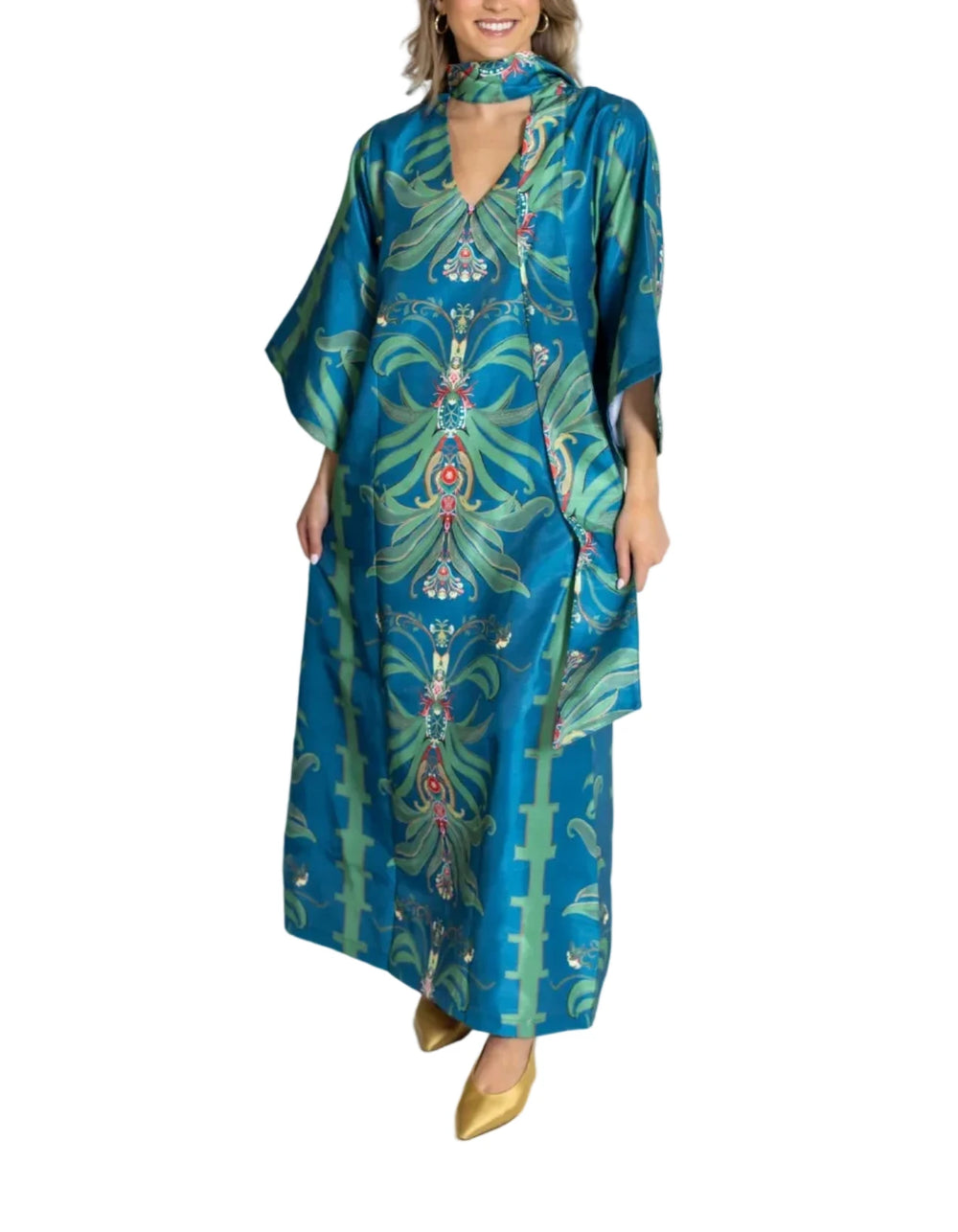 A woman modeling a long teal blue kaftan dress with a green and red botanical pattern, styled with a matching neck scarf and gold pointed shoes against a white studio background.