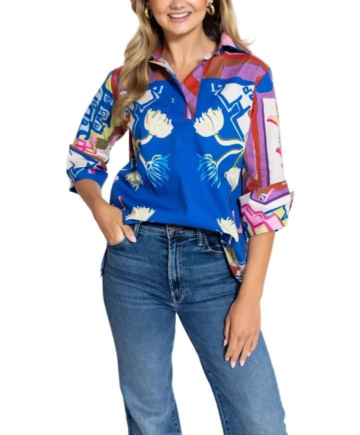 A woman modeling a vibrant blue collared shirt with a multicolor abstract pattern and white floral motifs, paired with blue jeans, against a plain white studio background.