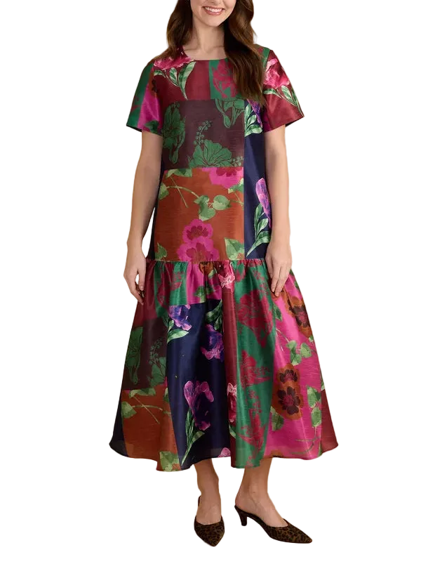 A woman modeling a short-sleeve midi dress with a vibrant floral print in pink, green, and navy blue, set against a solid tan studio background.