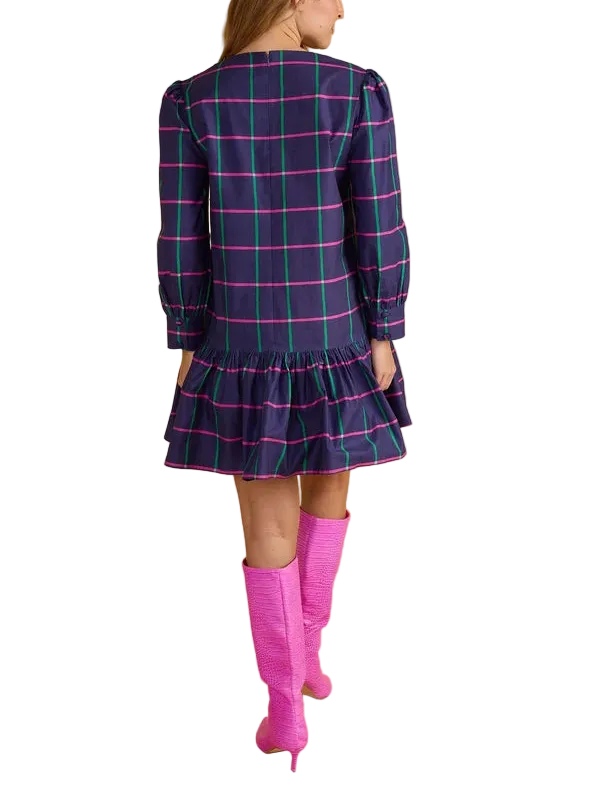 A woman shown from the back wearing a navy blue plaid mini dress with bright pink and green lines, paired with bright pink knee-high boots, against a solid beige studio background.