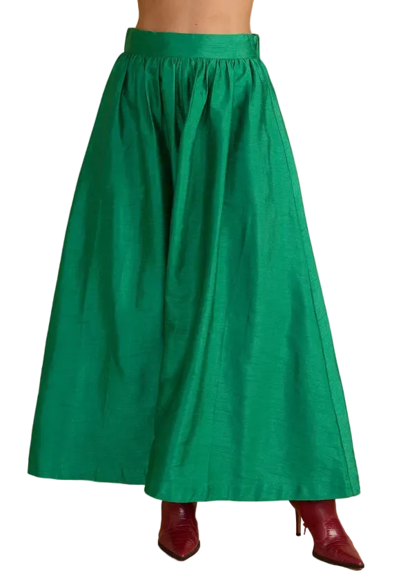 A bright green full-length skirt shown on a model from the waist down, paired with dark red pointed-toe heels, against a solid beige studio background.