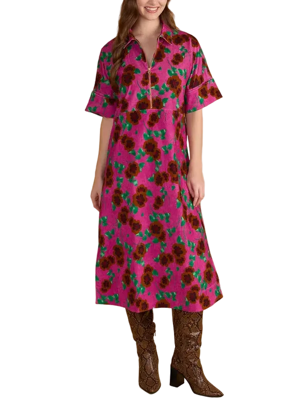A woman modeling a short-sleeve midi dress with a vibrant pink background and a dark brown and green floral pattern, paired with brown textured boots, against a solid beige studio backdrop.