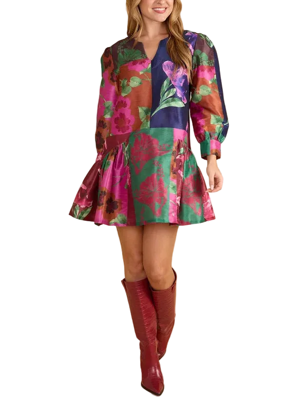 A woman modeling a short, long-sleeved mini dress with a vibrant multicolor floral patchwork print, paired with knee-high burgundy boots, against a solid beige studio background.