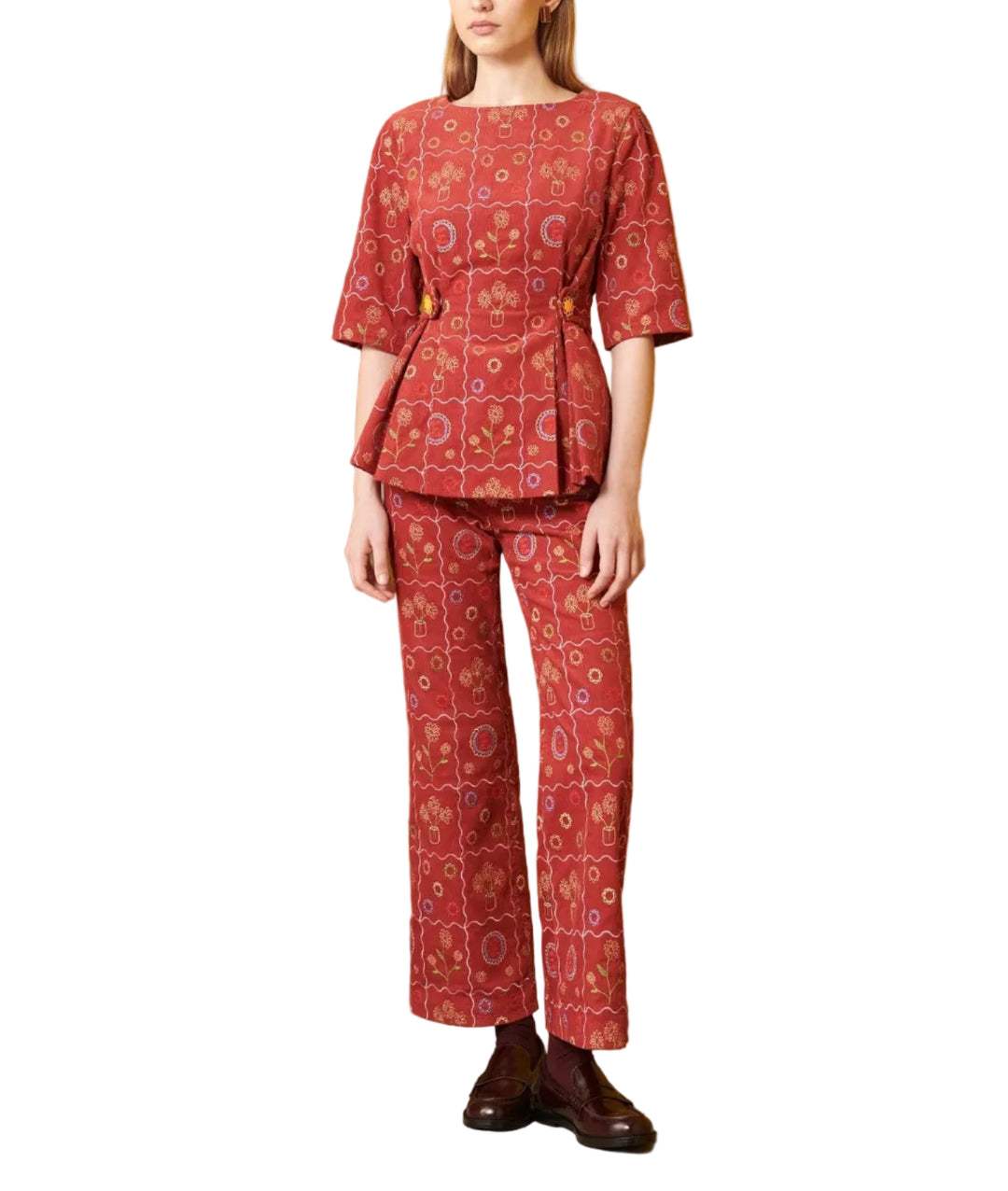 A model stands in a studio setting wearing a matching red patterned two-piece set consisting of a short-sleeve peplum top and wide-leg trousers. The set features a geometric and floral mandala pattern in red, orange, and white.