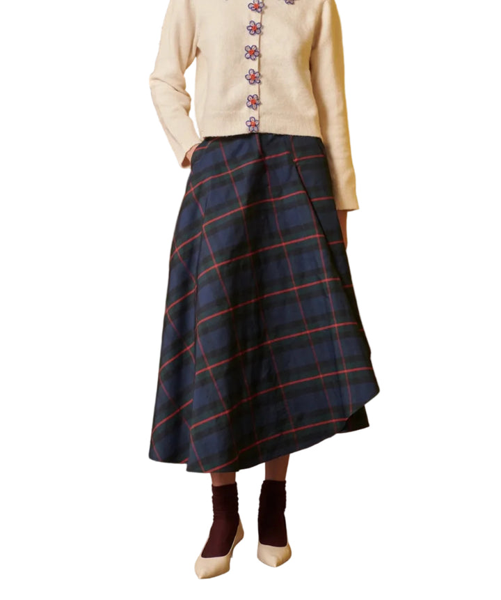 A model stands in a studio setting wearing a cream cardigan with a colorful patterned collar and buttons, paired with a navy blue plaid skirt, dark socks, and cream shoes. The background features a split design with a brown wood panel on the left and a solid mustard yellow on the right.