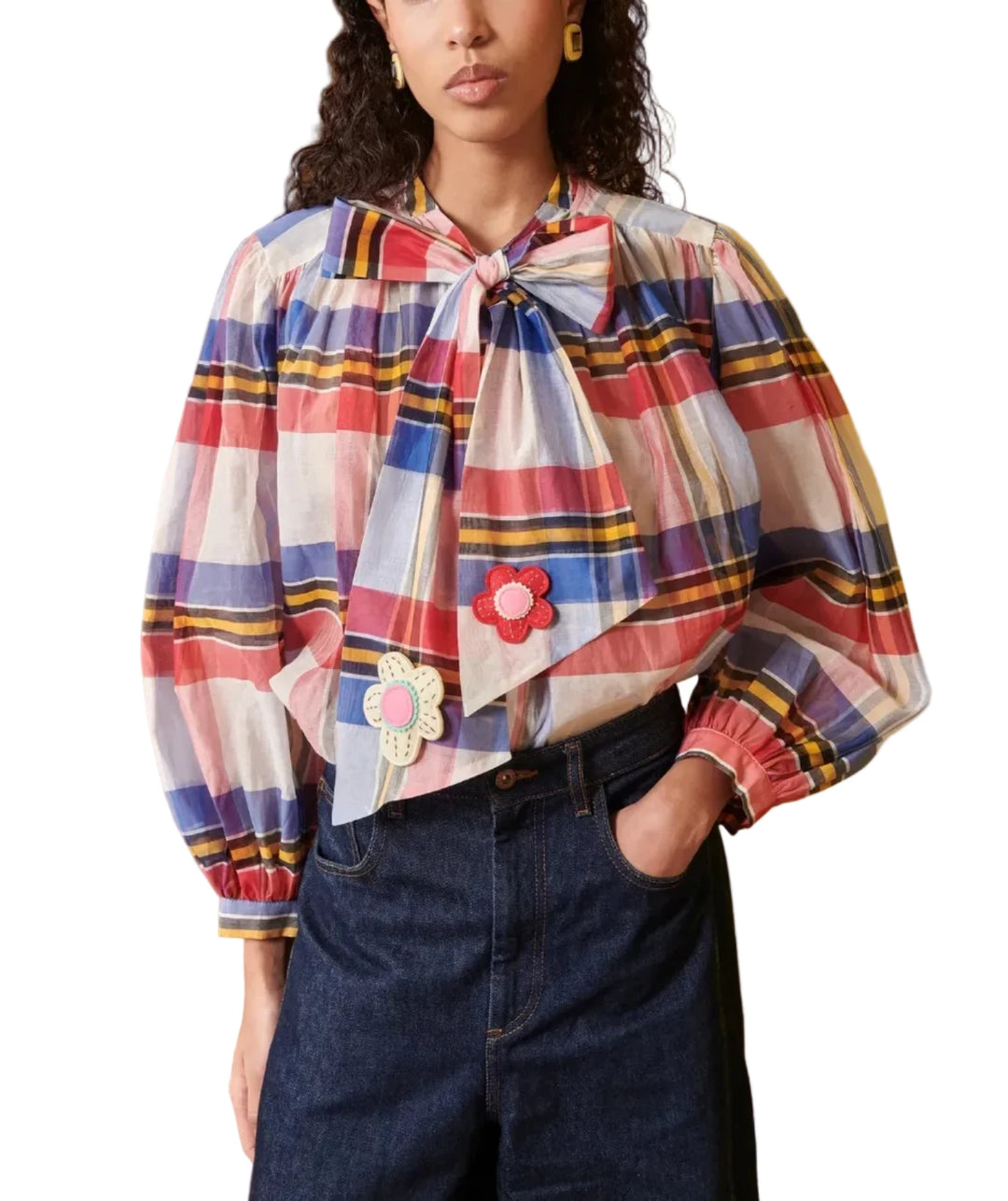 A model wearing a colorful plaid long-sleeve blouse with a large bow at the collar and embroidered flower details, paired with dark blue jeans, set against a split wood and yellow wall background.