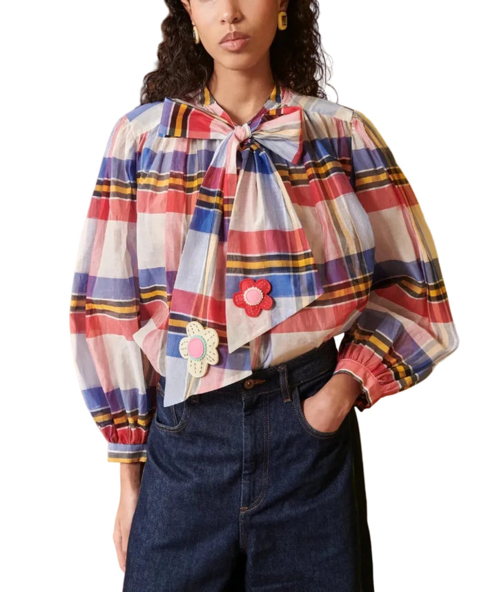 A model wearing a colorful plaid long-sleeve blouse with a large bow at the collar and embroidered flower details, paired with dark blue jeans, set against a split wood and yellow wall background.