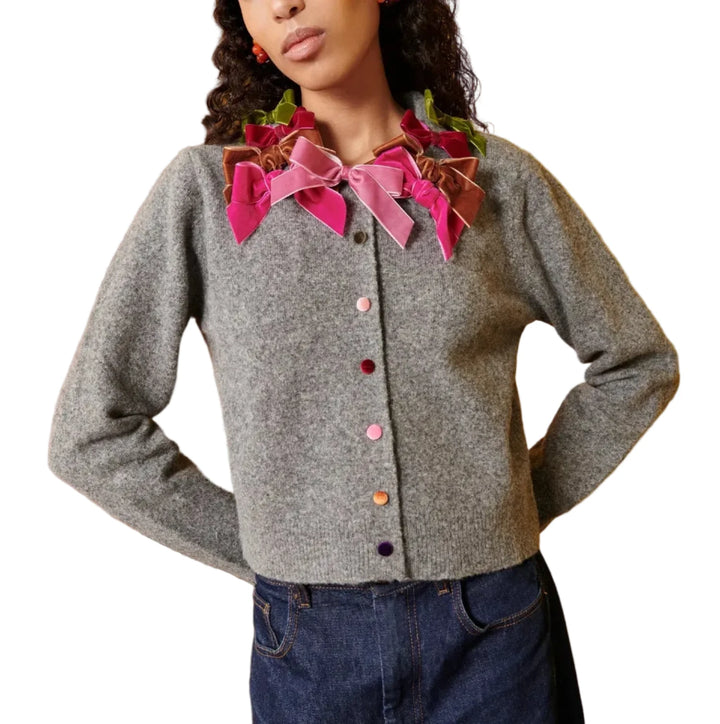 A model wearing a gray cropped cardigan with a large, decorative pink and magenta bow collar, paired with dark blue jeans, set against a vibrant yellow background.