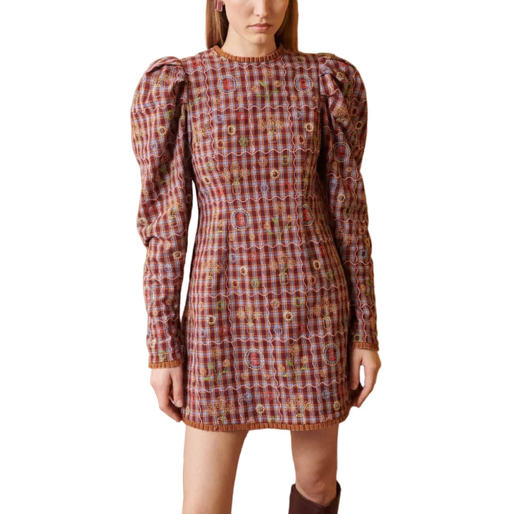 A model stands in a studio setting wearing a long-sleeved mini dress with a red, blue, and yellow plaid pattern and puff sleeves. The background features a split design with a textured light brown wood panel on the left and a solid mustard yellow wall on the right.