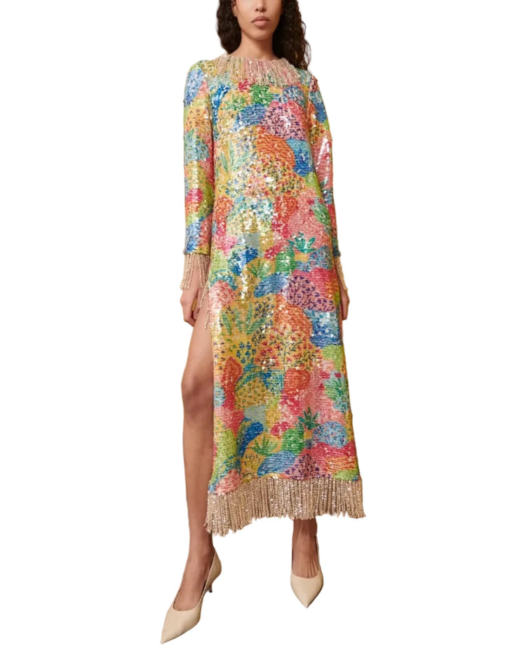 A model stands in a room with a mustard yellow wall, wearing a long-sleeved, high-slit midi dress covered in a dense multicolor floral and abstract print with a fringed hem. She is wearing beige pointed-toe heels.