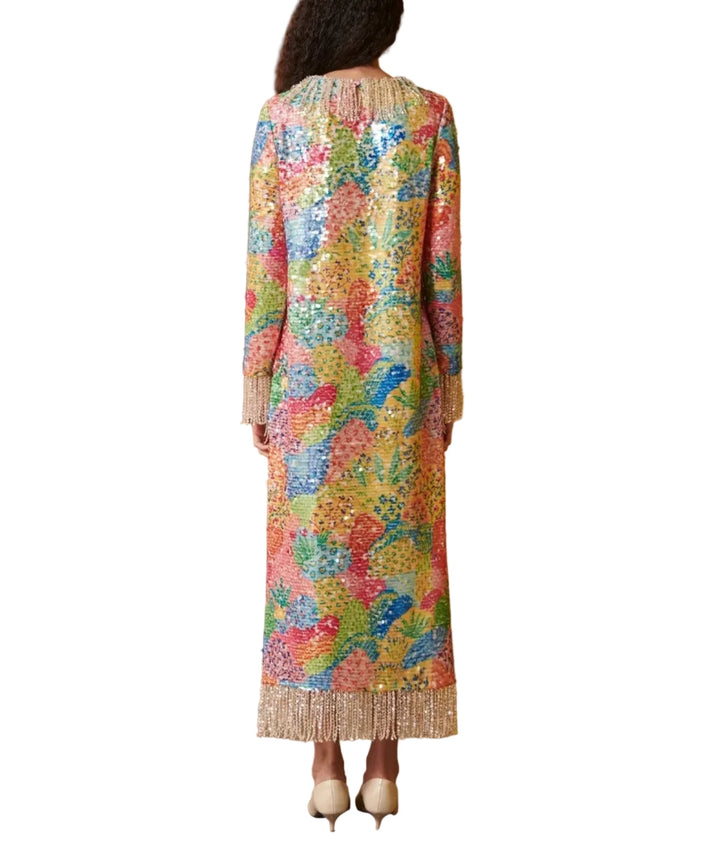 A woman stands with her back to the camera wearing a long-sleeved, fringed maxi dress covered in colorful floral sequins. The dress features a vibrant mix of yellow, pink, blue, green, and orange.
