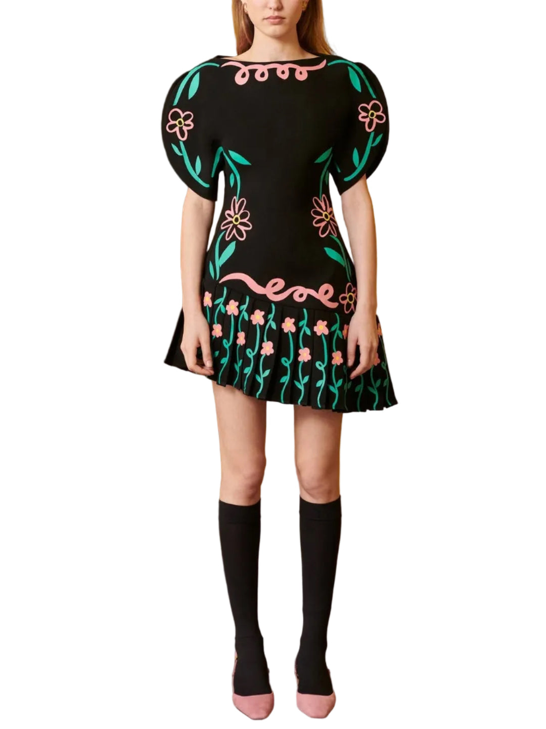 A model stands against a mustard yellow wall and wood paneling, wearing a black mini dress with colorful floral embroidery, black knee-high socks, and pink shoes.