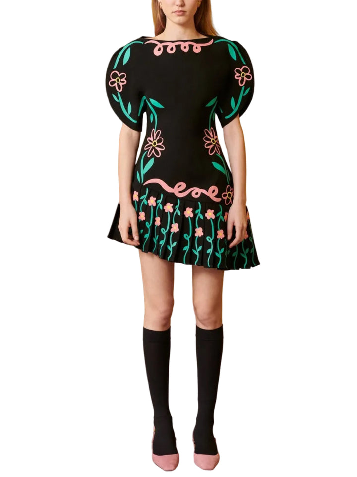 A model stands against a mustard yellow wall and wood paneling, wearing a black mini dress with colorful floral embroidery, black knee-high socks, and pink shoes.