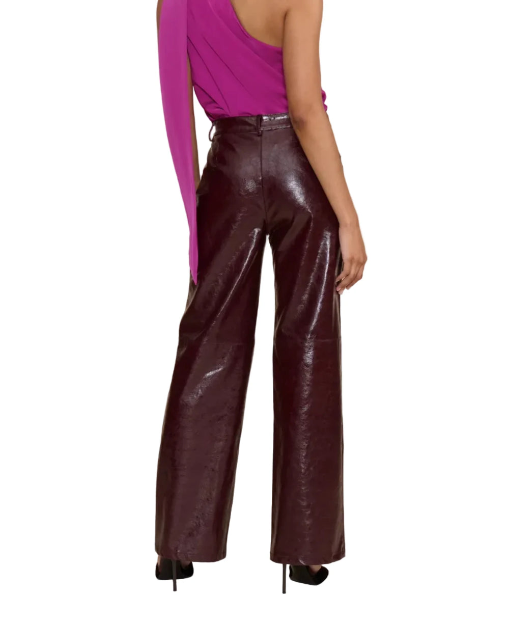 A woman shown from the back wearing a bright magenta one-shoulder draped top and shiny dark burgundy wide-leg pants, standing against a plain beige studio background.