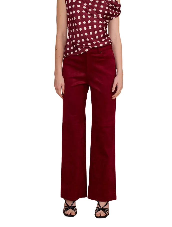 A model stands against a solid beige studio background wearing a burgundy one-shoulder top with white polka dots and matching burgundy wide-leg trousers.