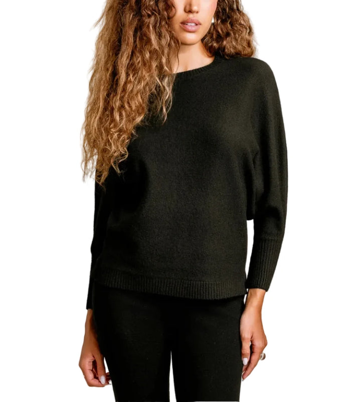 A woman with long, curly light brown hair wearing a dark olive green knit sweater and black pants, standing against a plain beige studio background.