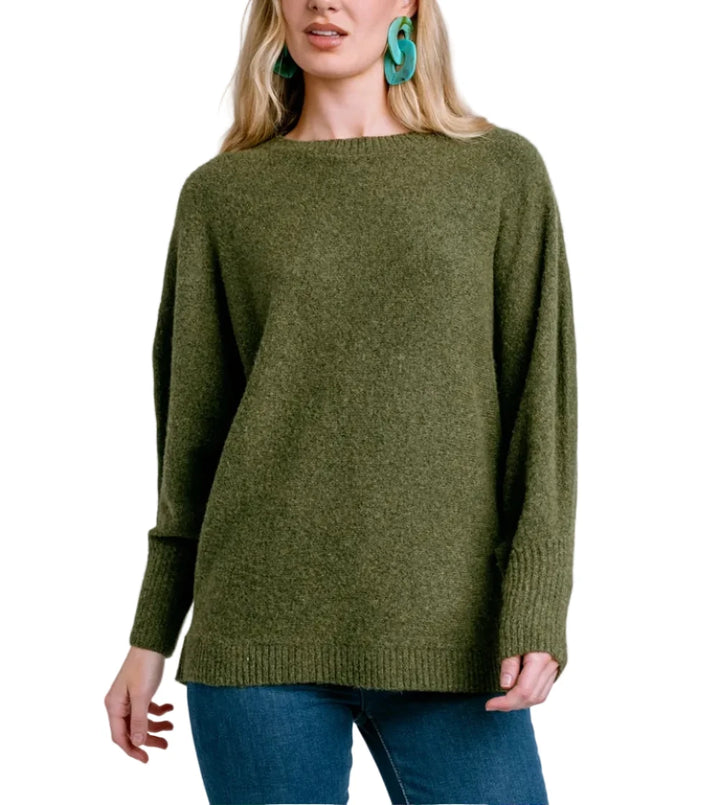 A woman modeling an oversized olive green knit sweater with a wide boat neckline, paired with blue jeans against a light blue studio background.