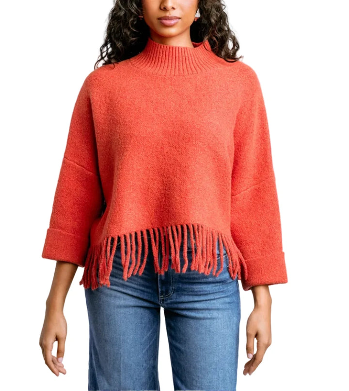 A woman wearing a coral orange knit mock-neck sweater with fringe trim at the hem and three-quarter sleeves, paired with blue jeans, against a light gray studio background.