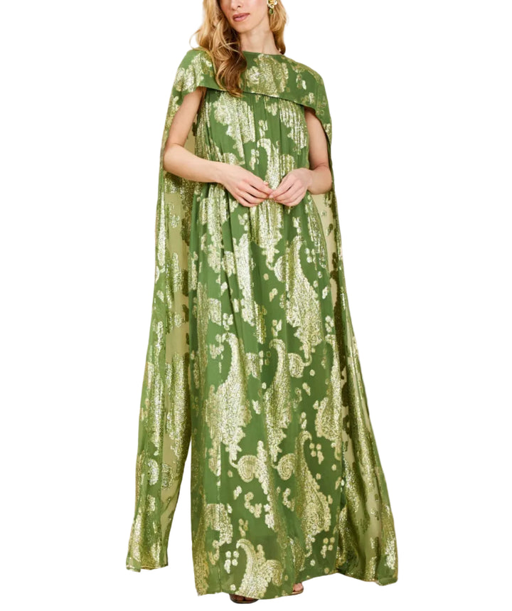 A woman modeling a floor-length green dress with a metallic gold paisley pattern and draped cape sleeves, set against a solid beige studio background.