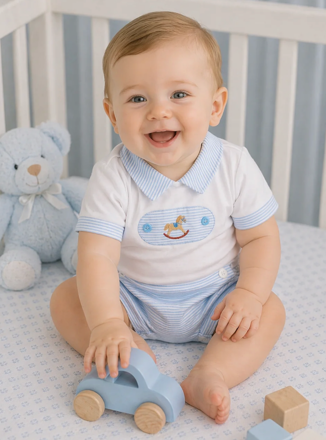A smiling baby sitting in a crib wearing a white short-sleeve romper with light blue stripes on the collar and sleeves, plus matching blue striped shorts. The baby is holding a light blue wooden toy car, with a blue teddy bear and small wooden blocks nearby.